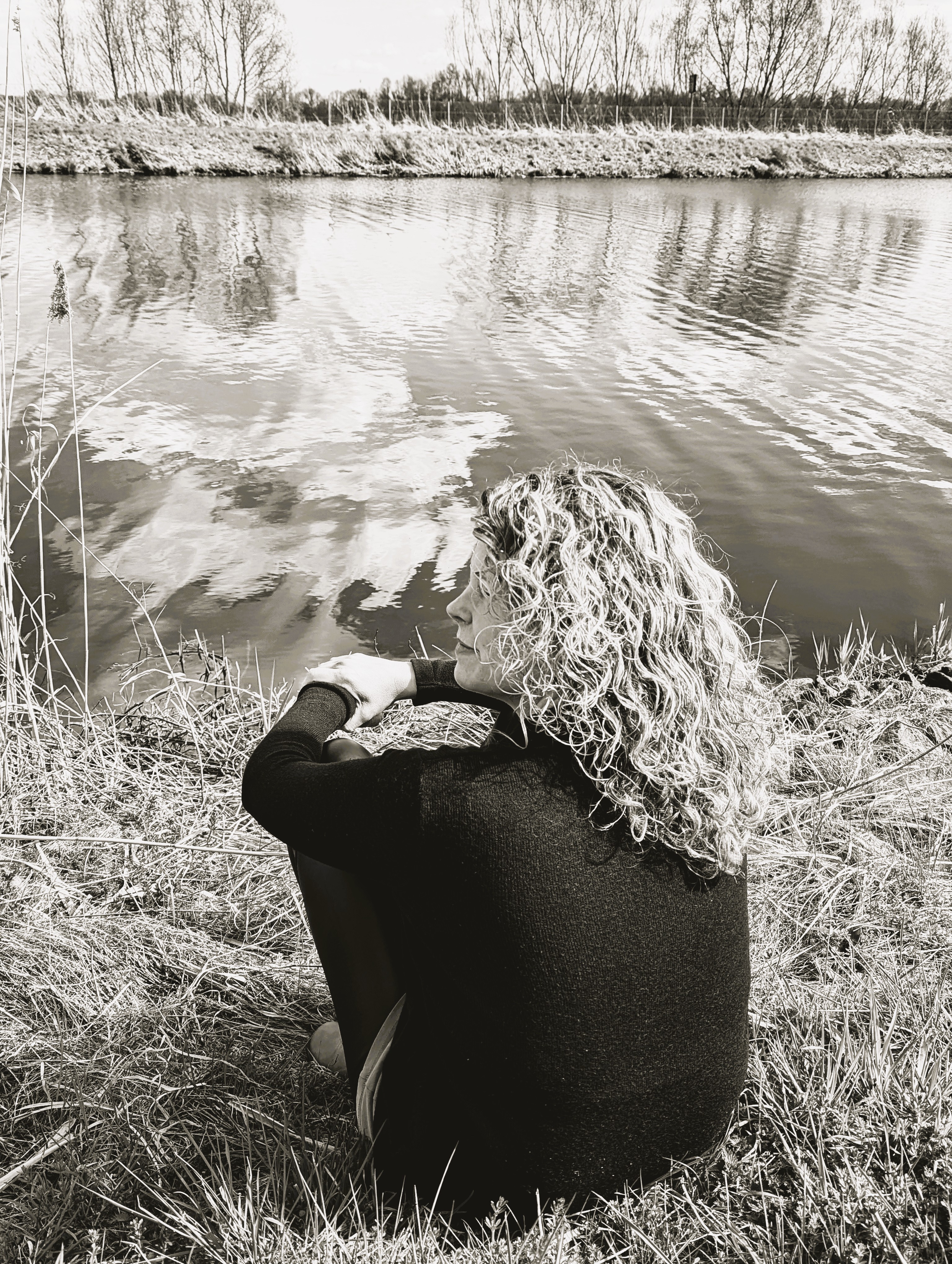 „Die Website-Inhaberin mit blonden Locken sitzt entspannt im Gras auf einer Wiese und guckt auf`s Wasser.“