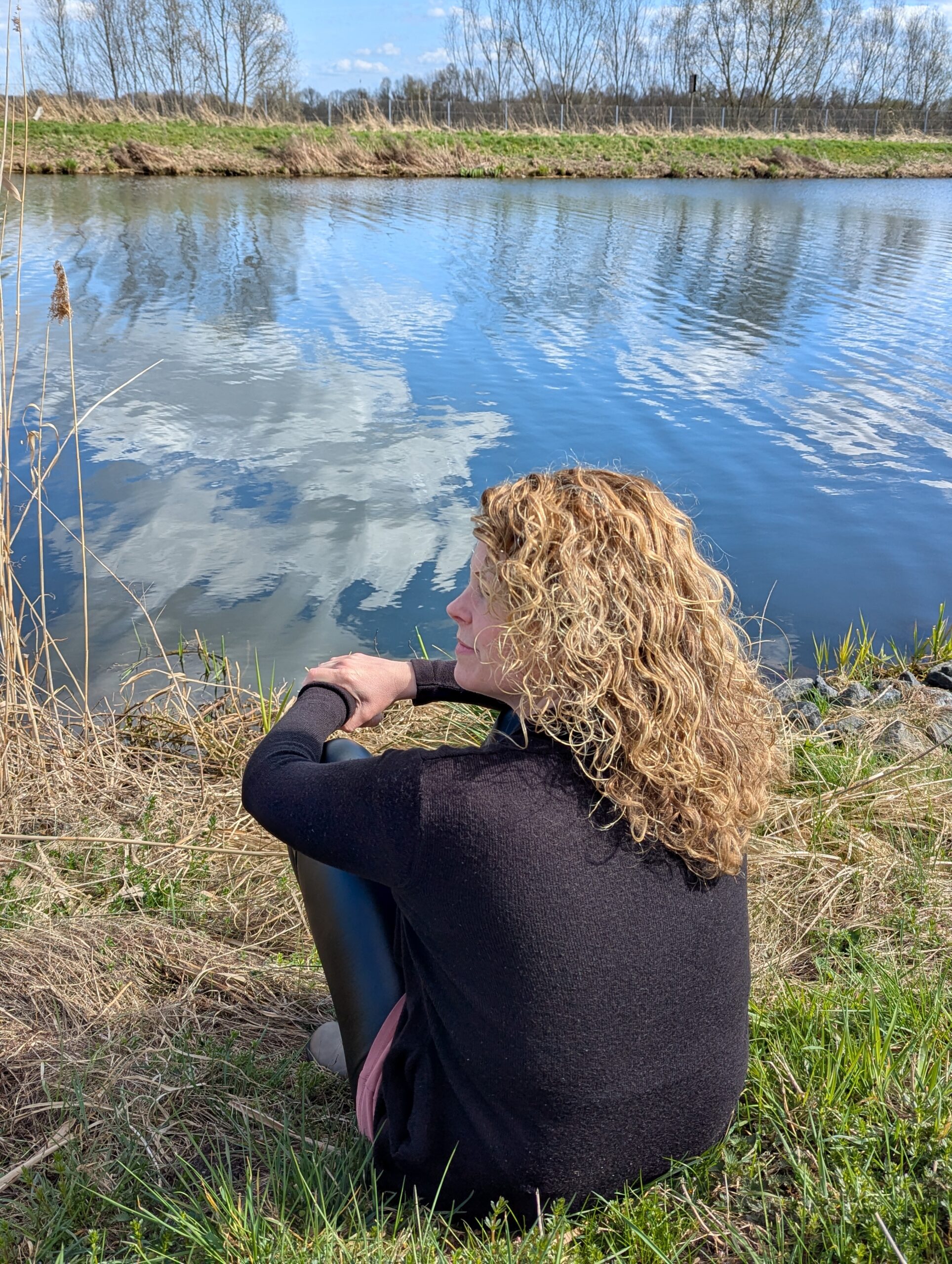 „Die Website-Inhaberin mit blonden Locken sitzt entspannt mit dem Rücken zu einem im Gras auf einer Wiese und guckt auf den Fluss.“